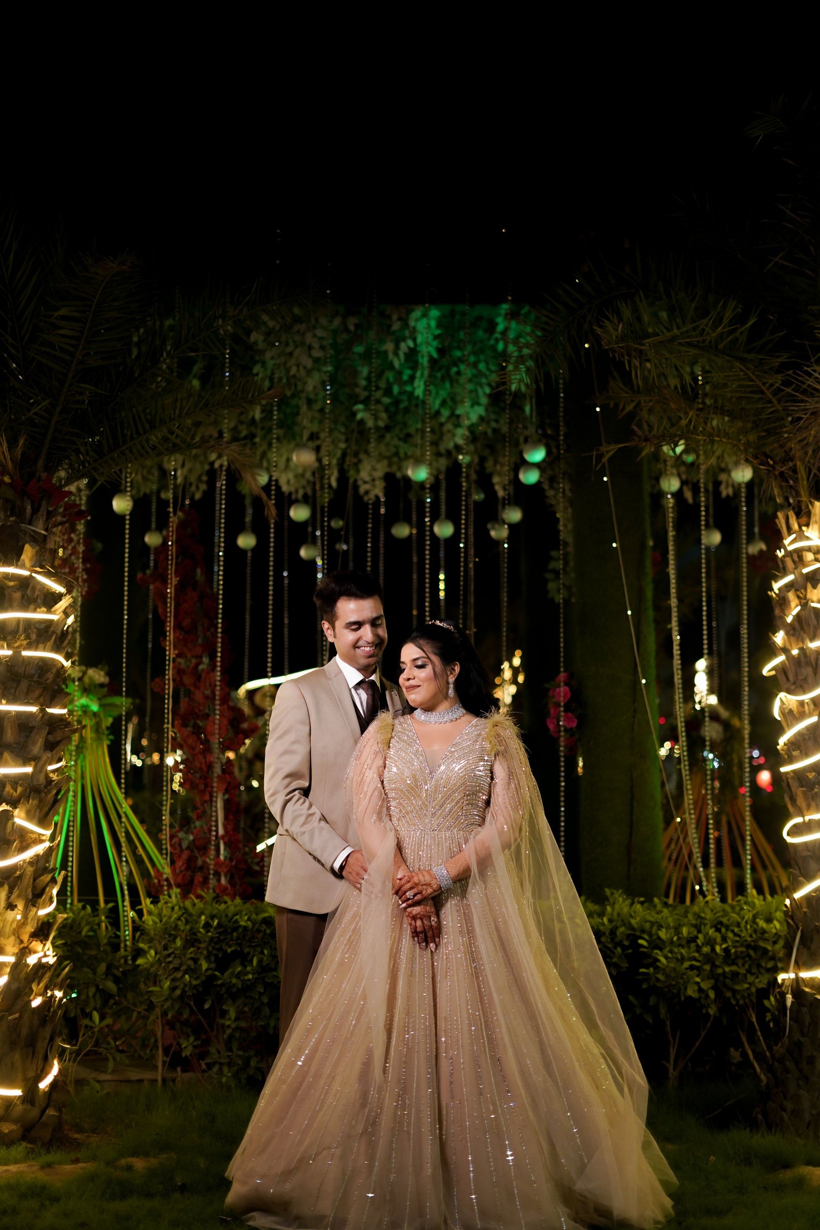 Sample stock photograph: An Indian wedding couple in champagne and tan attire stand under fairy lights; the bride wears a beaded gown, with henna on her hands.