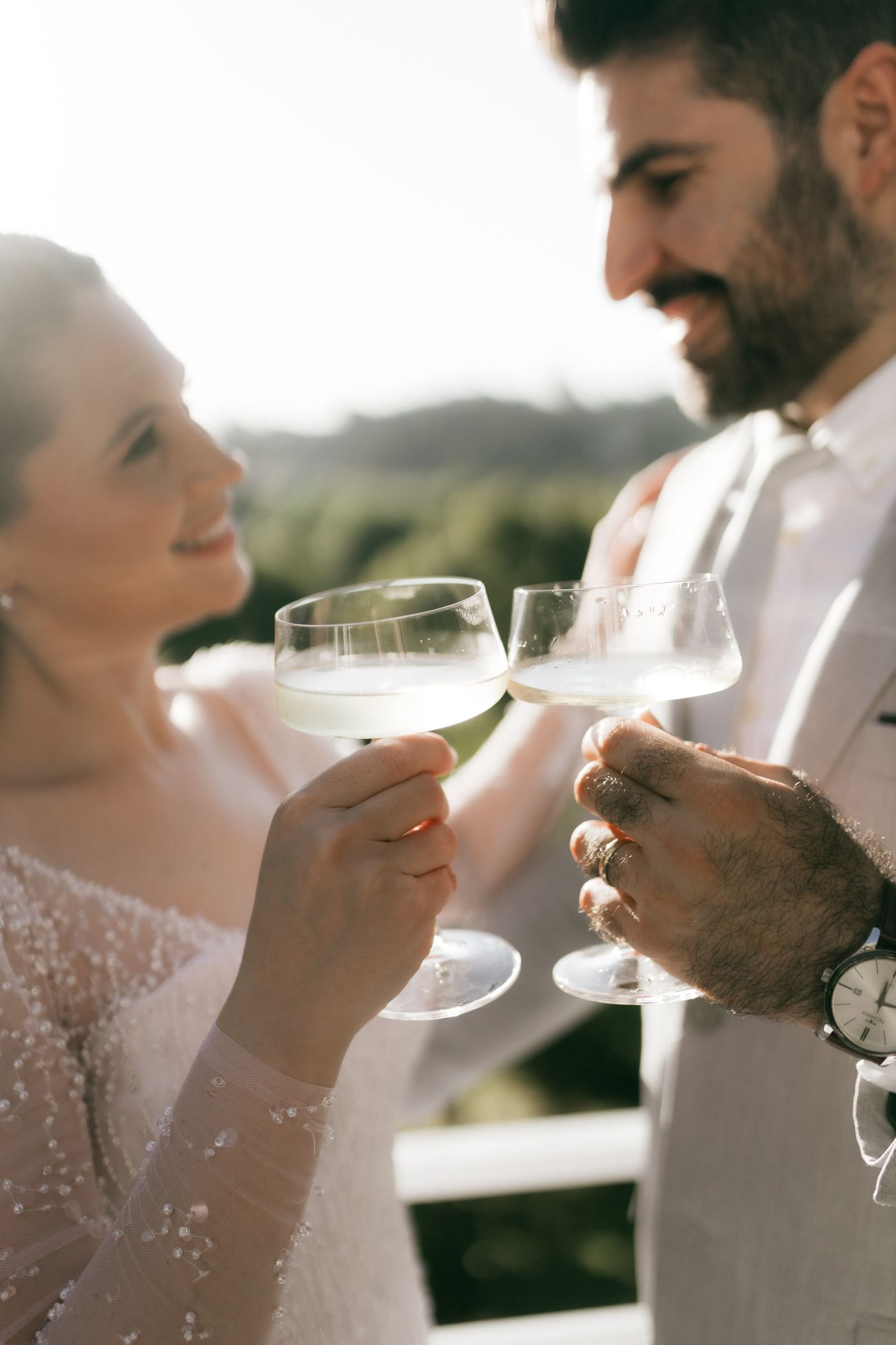 Sample stock photograph: Champagne coupes in the foreground while a couple in an ivory beaded gown and a light suit toast outdoors in golden light.